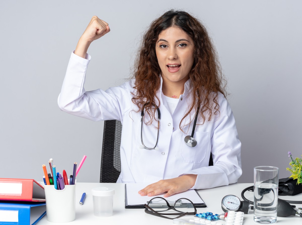 excited young female doctor wearing medical robe and stethoscope sitting at table with medical tools keeping hand on table looking at camera doing strong gesture isolated on white background